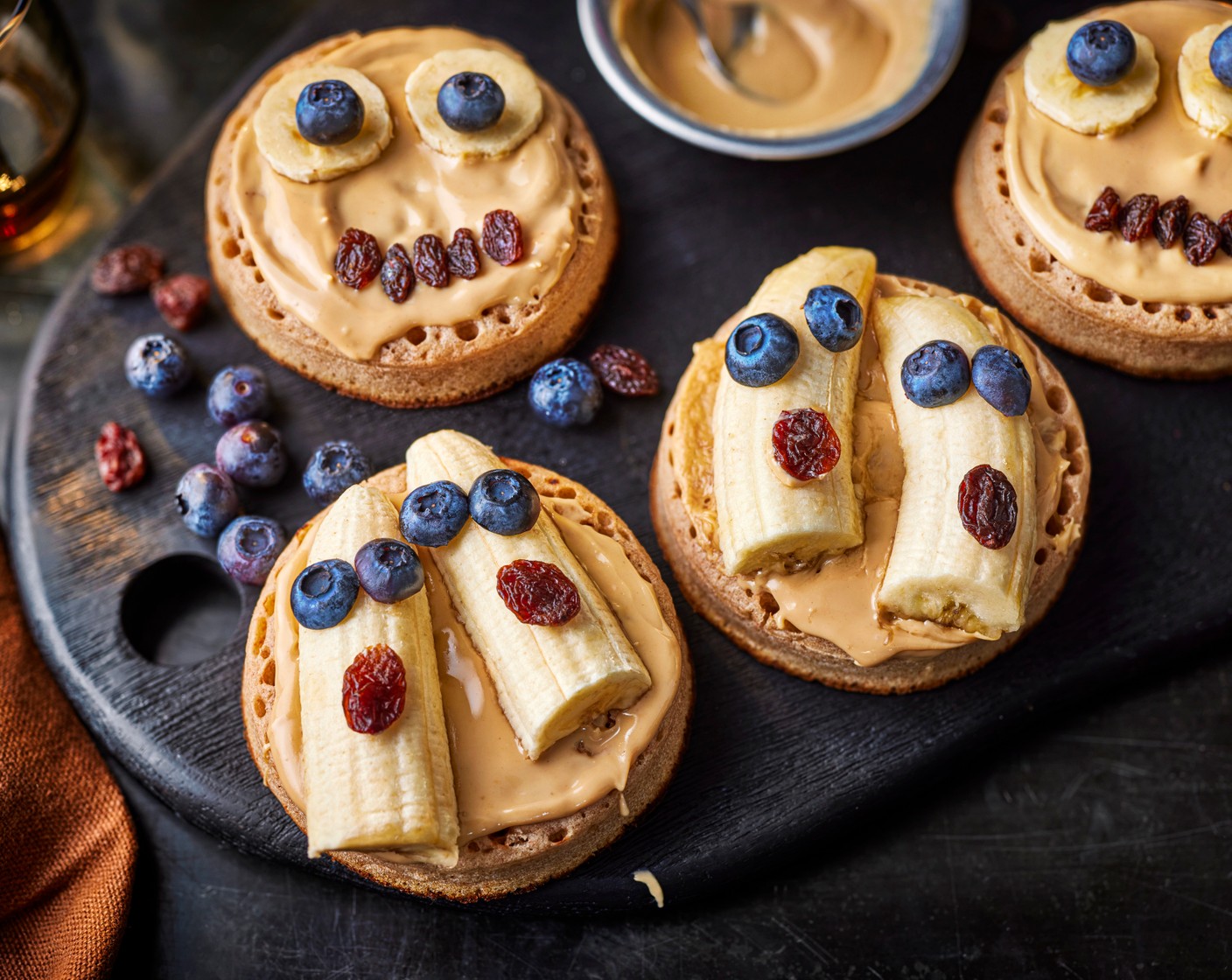 step 3 Place two slices of banana on top of each crumpet and top each with two blueberry halves to look like eyes, and a raisin (4) to look like a mouth (squish the raisins slightly to make them round).
