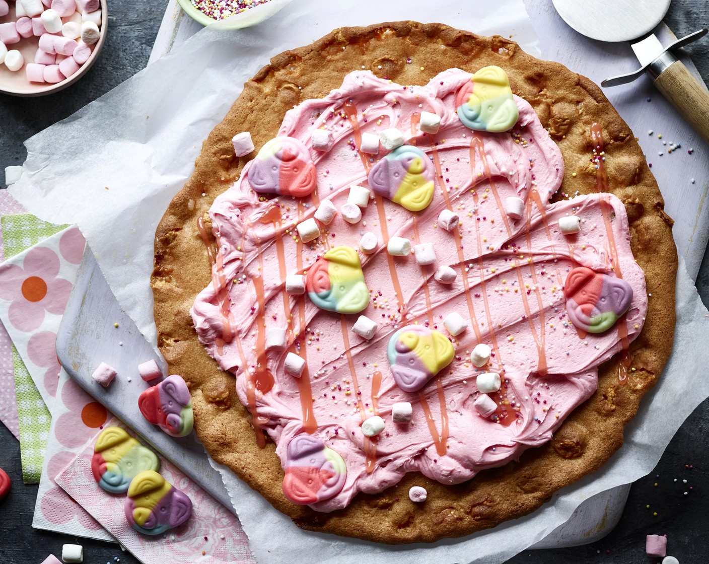 step 8 Spread the buttercream evenly onto the fully cooled cookie base, leaving some space around the edge, then drizzle the pizza with M&S strawberry sauce (to taste) before decorating with mini marshmallows (to taste), Percy Pig sweets (to taste) and jazzy sprinkles (to taste).